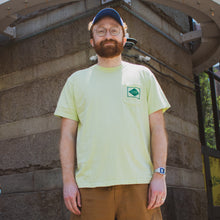 Load image into Gallery viewer, Man wearing a light yellow t-shirt with a logo, standing against a stone wall.