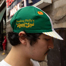 Load image into Gallery viewer, A man in profile wearing a green and white baseball cap with yellow embroidered text reading 'Feeding Philly's Heart Soul' and a small red and yellow logo patch on the side, photographed outdoors on a city street.