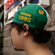 Load image into Gallery viewer, A man shown in profile wearing a green baseball cap with yellow text reading 'since 1893' and a small red-and-yellow crest patch. The cap has white side panels and striped brim details, photographed outdoors in front of a storefront.
