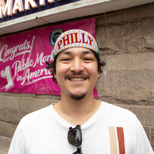Load image into Gallery viewer, A smiling man wearing a white t-shirt and a white striped baseball cap with 'PHILLY' in red letters, standing in front of a stone wall and a pink banner.
